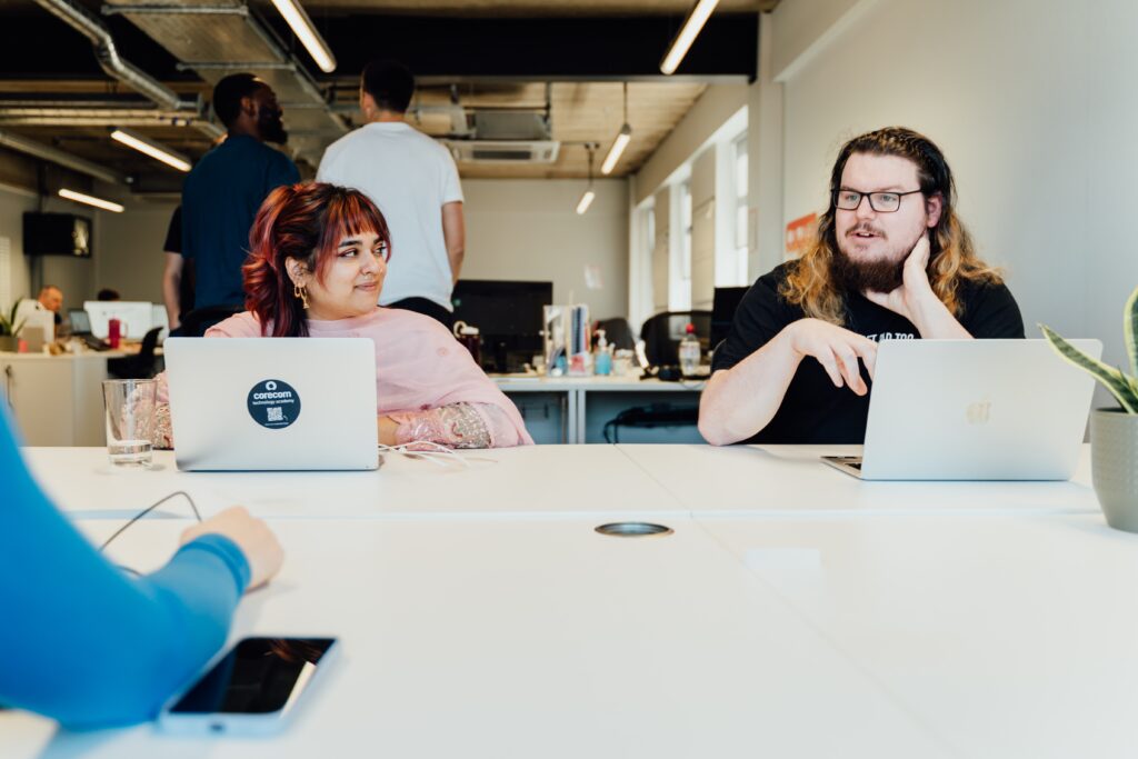 person with red and black hair on the left in a pink top with a CoreCom HP laptop looking at person to their right with brunette ombre blonde hair explaining something
