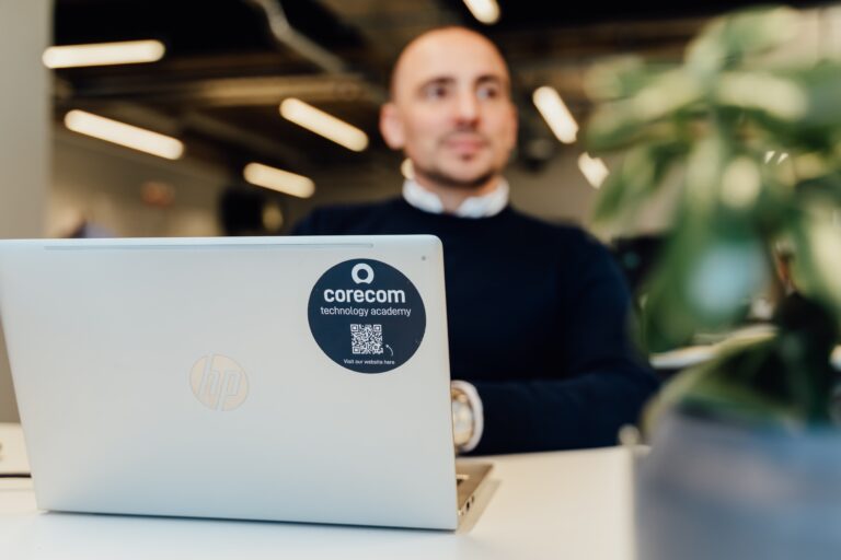 In focus is a silver HP laptop with a blue circle QR code sticker, out of focus is James in the background